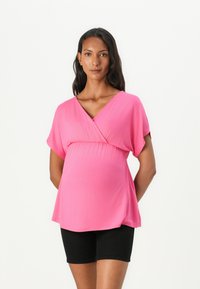Rosa graviditetstopp med V-hals og korte ermer, med en rynket søm under brystet. Kombinert med svarte shorts. Mykt stoff, avslappet passform.
