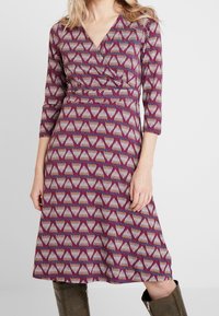 V-Ausschnitt, drei Viertel Ärmel Kleid in Lila mit einem geometrischen Dreiecksmuster in Grau und multikulturellen Akzenten, mit einer gerafften Taille.