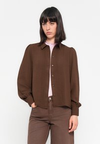Uma mulher com cabelo liso e escuro usa uma blusa castanha de botão sobre uma blusa de gola plissada rosa clara e calças castanhas em combinação, com as mãos nos bolsos.
