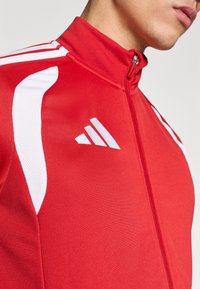 Κόκκινο αθλητικό μπουφάν με λευκές λεπτομέρ ειες, ψηλό γιακά και κλείσιμο με φερμουάρ. Διαθέτει υφή υφάσματος και έμβλημα λογότυπου στο στήθος.