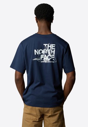Osoba ubrana w granatowy T-shirt z białą grafiką gór i napisem „THE NORTH FACE Never Stop Exploring” na plecach, widoczne jasnobrązowe spodnie.