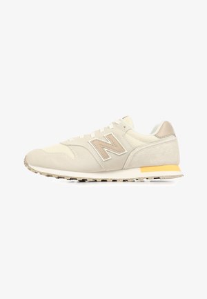 Zapatilla New Balance beige con paneles de ante y malla, logo "N" beige, entresuela blanca y detalle amarillo en el talón.