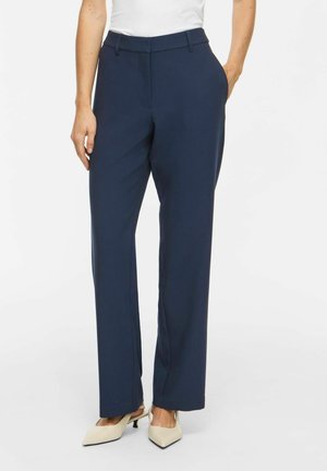 Femme portant un pantalon droit bleu marine avec des poches latérales, assorti à des talons bas blancs à bout pointu et un haut blanc.