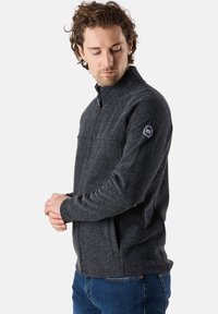 Schwarze, melierte Zip-Jacke mit hohem Kragen, strukturiertem Stoff, taillierter Passform und Logopatch auf dem linken Ärmel.