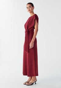 Punainen polvipituinen mekko, jossa on korotettu vyötärö, lyhyet hihat ja sileä pinta. Yhdistetty mustiin, matalakorkoisiin sandaaleihin. Minimalistinen muotoilu.