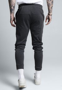 Person med tatuerade armar har på sig mörkgrå joggingbyxor, vita ankelsockor och vita Adidas-sneakers, står mot en enkel grå bakgrund.