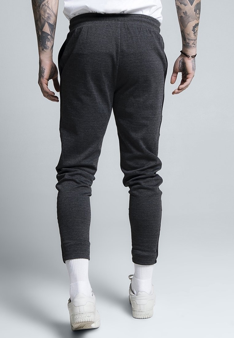 Person med tatuerade armar har på sig mörkgrå joggingbyxor, vita ankelsockor och vita Adidas-sneakers, står mot en enkel grå bakgrund.