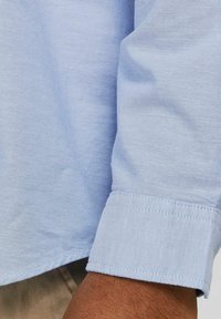 Camisa de manga larga azul claro con tela texturizada; detalle de puño visible con costura prolija; brazo sosteniendo la camisa cerca de una tela beige abajo.
