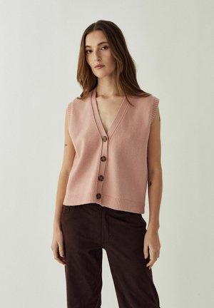 Donna che indossa un gilet rosa senza maniche con bottoni e pantaloni marrone scuro, in piedi contro uno sfondo chiaro e semplice.