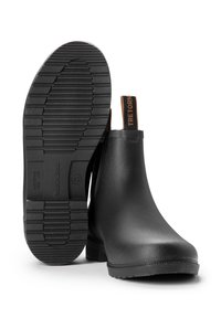 Musta kuminen nilkkasaappaat, joissa on joustavat sivupaneelit, teksturoitu pohja ja ruskea logotunnus. Yksinkertainen muotoilu, kestävä rakenne ja vedenpitävä materiaali.