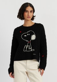 Pull en maille noire avec un design Snoopy brodé, col rouge et petits accents de cœur rouge sur les manches.