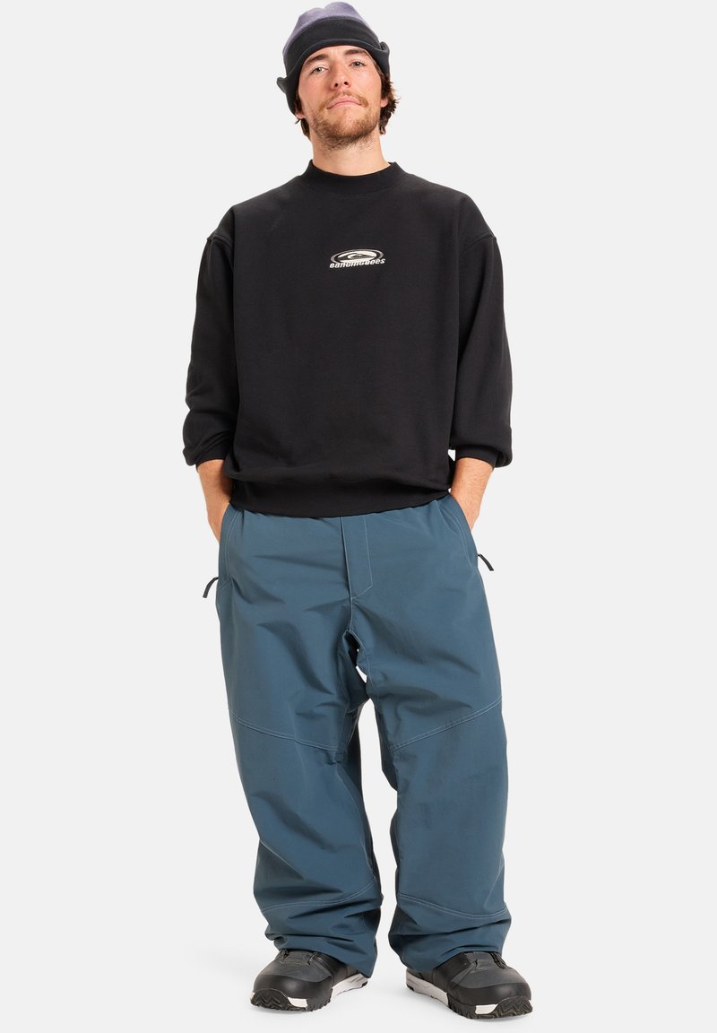 Zwart sweatshirt met logo, losse blauwe broek en zwarte sneeuwlaarzen. De outfit heeft een casual, praktische uitstraling voor buitenactiviteiten.