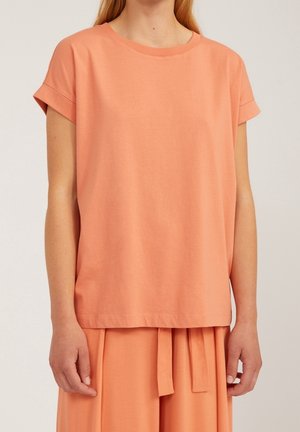 Kobieta ubrana w luźną, koralowo-pomarańczową bluzkę z krótkim rękawem oraz dopasowaną spódnicę z wiązanym paskiem w talii.