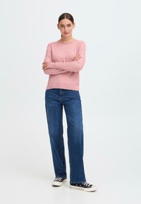 Lys rosa strikket genser med ribbestruktur, kombinert med blå rette jeans. Modellen har på seg svart og hvite joggesko, stående med armene krysset.