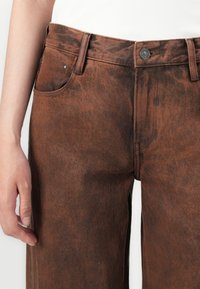 Brown denim trousers with a textured surface, front pockets, and a metal button at the waist. Features a high-rise fit and straight legs.