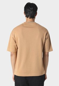 Muž s krátkými tmavými vlasy, oblečený v jednoduchém béžovém tričku s krátkým rukávem a černých kalhotách, pohledem zezadu na světle šedém pozadí.