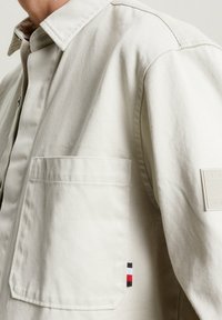 Vaaleanbeige puuvillasta valmistettu takki, jossa on nappikiinnitys edessä, rintatasku ja hihassa logo-patch punaisilla, valkoisilla ja sinisillä yksityiskohdilla.