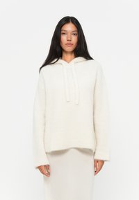 Felpa bianca con texture, dalla vestibilità ampia, con polsini a coste e lacci sul collo. Indossata sopra un indumento leggero e fluttuante.