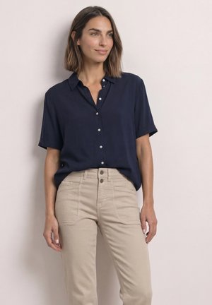Vrouw met schouderlang bruin haar, gekleed in een marineblauw kortemouwen overhemd dat in een beige broek is gestopt, staand tegen een effen achtergrond.