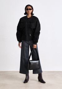 Black cropped bomber jacket with two front pockets, paired with wide-leg dark denim jeans. Model carries a small black handbag and wears black boots.