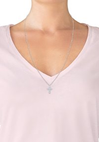 Silberne Kettenhalskette mit einem Kreuzanhänger, der mit kleinen klaren Steinen geschmückt ist, liegt auf einem hellrosa V-Ausschnitt-Shirt.