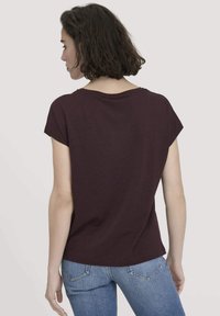 Frau mit schulterlangen dunklen Haaren, die ein kurzärmeliges, dunkelviolettes T-Shirt und Bluejeans trägt und sich von einem schlichten Hintergrund abwendet.