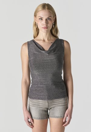 Mujer rubia con top sin mangas, brillante en gris oscuro con escote drapeado y shorts de mezclilla gris claro sobre fondo liso.