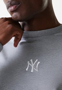 Grå sweatshirt lavet af blødt stof, der har en ribbet krave og et hvidt broderet logo på brystet. Nærbillede af teksturen.