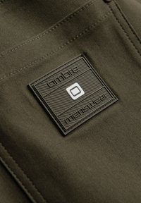 Étiquette carrée en tissu noir avec « Ombre » et « Menswear » embossés sur un tissu cousu de couleur vert olive.