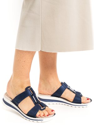 Füße in marineblauen Sandalen mit offenem Zehenbereich und weißen Sohlen, kombiniert mit beigen, verkürzten Hosen, auf weißem Hintergrund.