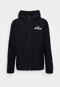Černá kapucí bunda s předním zipem, na levé straně hrudi s bílým logem "Hollister" a bočními kapsami. Hladká nylonová textura.