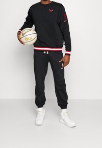 Sudadera negra con dobladillo de rayas rojas y blancas, con un logotipo en el pecho; pantalones jogger negros a juego; zapatillas blancas; sosteniendo un balón de baloncesto.