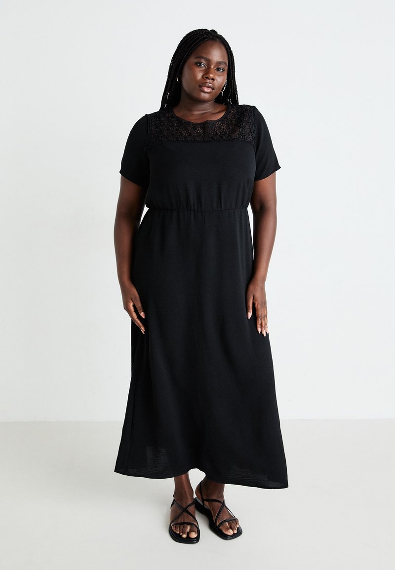 Vestido negro maxi con mangas cortas y un panel de encaje en el escote. Presenta una cintura elástica y una falda fluida. Sandalias negras de tiras.