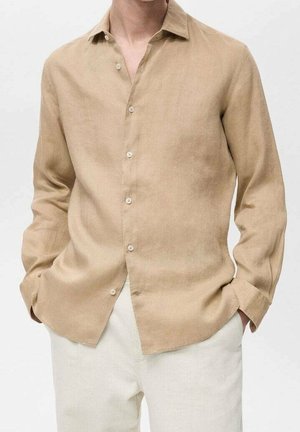 Homme portant une chemise beige à manches longues boutonnée avec col et un pantalon blanc, mains dans les poches sur un fond uni.