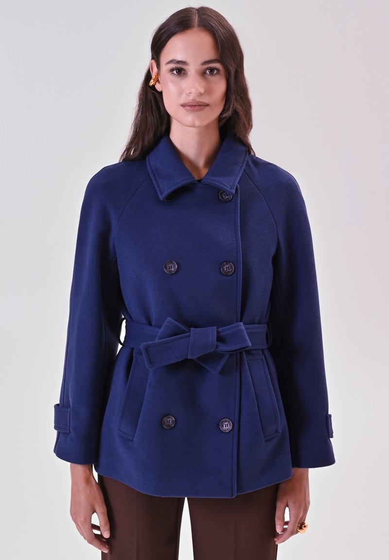 Cappotto doppiopetto blu navy con vita cinturata, colletto strutturato e tessuto textured. Presenta quattro bottoni e dettagli sui polsini.