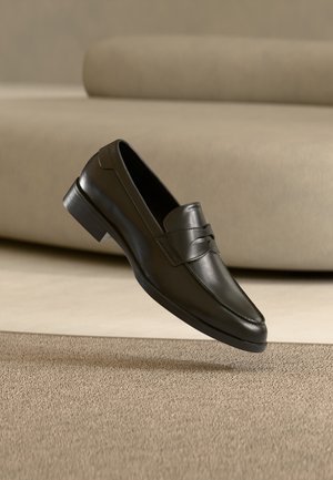 Mocassin en cuir noir flottant au-dessus d'un tapis beige avec un canapé beige en arrière-plan, mettant en valeur le design classique pour hommes sans lacets.
