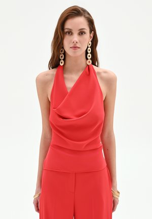 Giovane donna che indossa un top rosso drappeggiato senza maniche e pantaloni abbinati, accessoriata con lunghi orecchini circolari dorati e braccialetti d'oro.