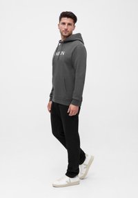 Grijze hoodie met een voorzak en logo, gecombineerd met zwarte broek en witte sneakers. Model staat tegen een effen achtergrond.