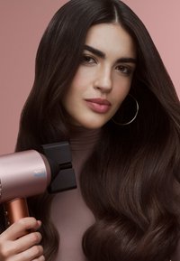 Asciugacapelli Dyson in rosa metallico con un beccuccio nero, caratterizzato da un design elegante, impugnatura ergonomica e attaccato a capelli lunghi, lucenti e scuri.