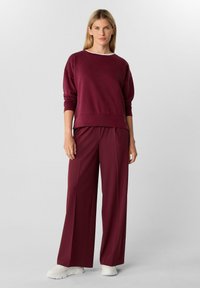 Vrouw die staat in een bordeauxlange mouw top en bijpassende wijde broek met witte casual sneakers tegen een neutrale achtergrond.