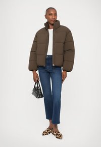 Chaqueta de plumón marrón con cuello alto, combinada con jeans azules ajustados y zapatillas de leopardo. Los accesorios incluyen un pequeño bolso negro.
