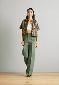 Chemise à imprimé serpent raccourcie, crop top blanc et pantalon vert taille haute. Fond en parquet en bois, mur gris clair. Texture lisse.