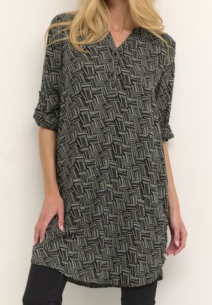 Femme portant une longue tunique à motifs noir et blanc avec des manches retroussées, associée à un pantalon noir.