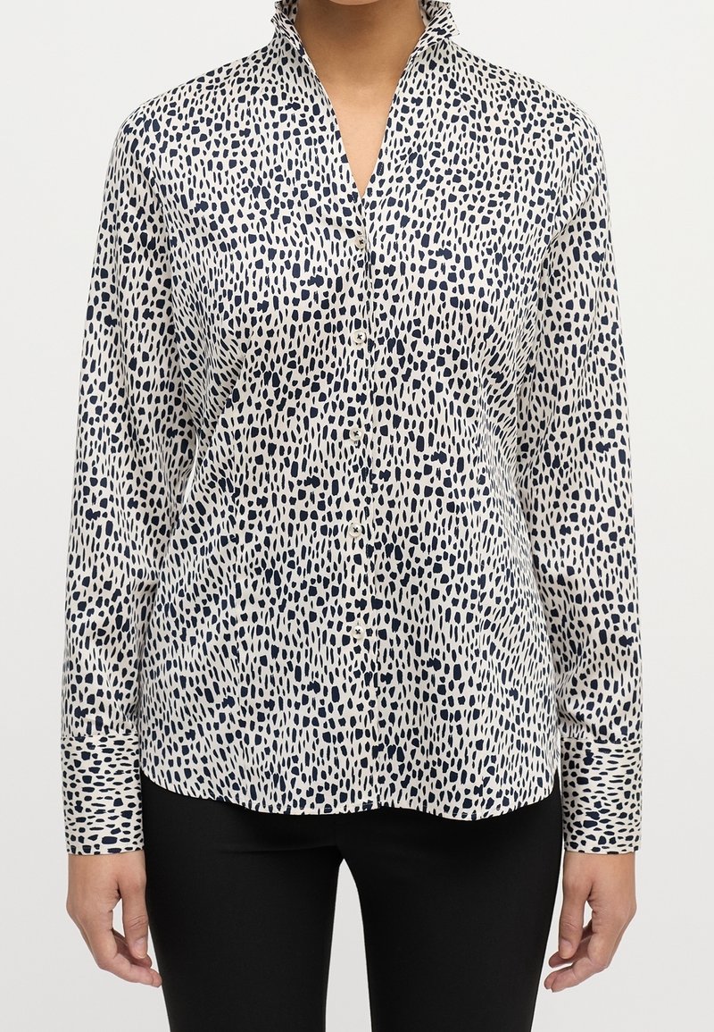 Blouse blanche avec un motif abstrait bleu marine, à manches longues et col montant. Fabriquée en tissu léger, elle dispose d'une fermeture à boutons sur le devant.