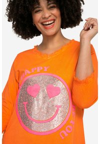 Oranges Baumwoll-T-Shirt mit ausgefranstem Ausschnitt und Ärmeln. Es zeigt ein silberglänzendes Smiley-Gesicht mit pinken Herzen als Augen und dem Wort "HAPPY."