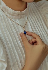 Collier en chaîne dorée avec une pierre bleue en forme de poire entourée de petites pierres claires, porté sur un tissu texturé de couleur claire.