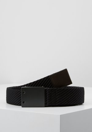 Ceinture en tissu tissé noir avec une boucle rectangulaire noire mate, enroulée et posée sur une surface blanche sur un fond gris clair.
