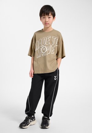 Chłopiec stoi z jedną ręką w kieszeni, ubrany w oversize beżowy T-shirt "Made to Move", czarne spodnie dresowe z białymi paskami i czarne trampki.