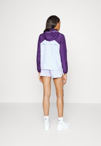 Chaqueta ligera en bloque de color en tonos púrpura y azul claro, con capucha; combinada con pantalones cortos a juego. Presenta un diseño elegante y acentos sutiles.