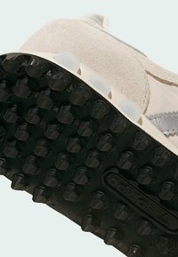 Αθλητικό παπούτσι με μπεζ επάνω μέρος από σουέτ και ύφασμα, μαύρη εξωτερική σόλα από υφή καουτσούκ, κυκλικά αντιολισθητικά καρφιά και ασημί διακοσμητική ρίγα.